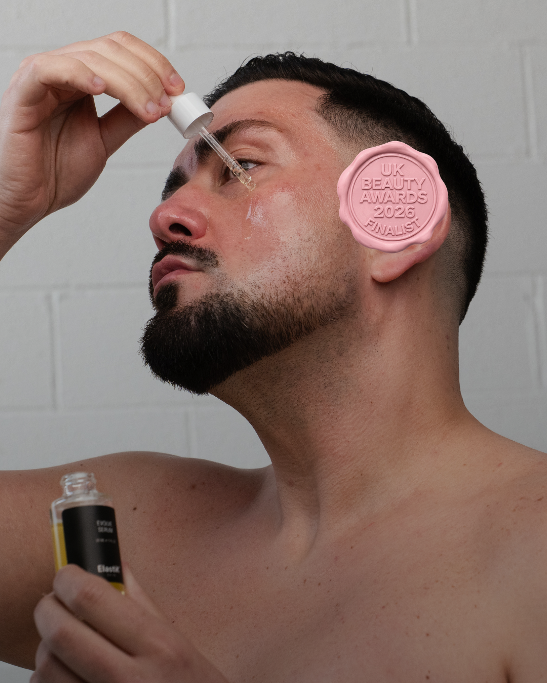 Man applying skincare product with a dropper, wearing a pink badge with text, against a white tiled wall.
